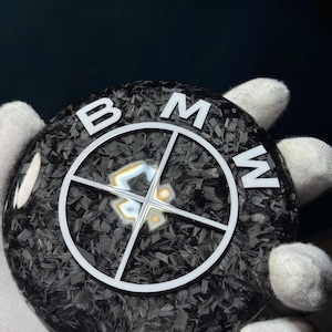 May include: A round, black carbon fiber emblem with the white letters "BMW" arranged around a white crosshair design. The emblem is held by a gloved hand, with a dark background.