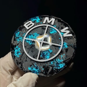 May include: A round, black carbon fiber emblem with the BMW logo in white. The emblem has a textured surface and is accented with bright blue glitter. The BMW logo is centered and features the letters "B", "M", and "W".