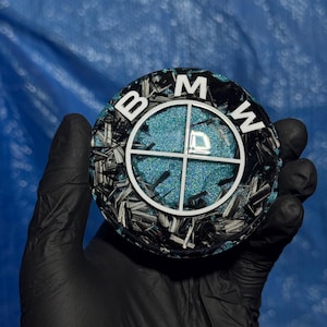 May include: A round emblem featuring the BMW logo, with the letters "B", "M", and "W" in white. The emblem has a black and silver carbon fiber pattern around a teal, glittery center. The emblem is held by a gloved hand.