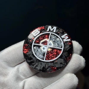 May include: A round emblem with the BMW logo. The emblem features a black carbon fiber base with red glitter accents. The BMW logo is white, with a silver and red segmented center.
