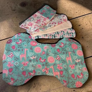 May include: A set of baby bibs and burp cloths with a floral and animal print. The bib is teal with pink giraffes, flowers, and butterflies. The burp cloths have coordinating patterns in pink, white, and teal. The items are made of soft fabric.