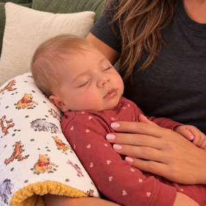 May include: A white nursing pillow featuring a repeating pattern of Winnie the Pooh characters including Tigger, Eeyore, and Piglet, with a textured mustard yellow underside, supporting a sleeping infant wearing a maroon long-sleeve shirt.