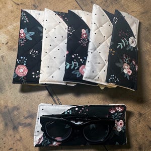 May include: Black sunglasses rest in a floral print case, with several matching cases above. The cases feature a quilted design with black fabric, white polka dots, and floral patterns in pink, white, and green.