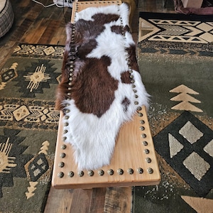 May include: A wooden bench with a cowhide seat. The cowhide is brown and white. The bench has brass-colored nailhead trim around the edges. The bench is on a wooden floor with a green rug.