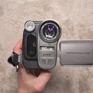 May include: A silver and black Sony Handycam camcorder with a lens that reads "Optical 20x". The screen reads "990x". The Handycam is held in a person's hand.