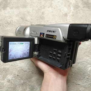 May include: A silver and black Sony camcorder with an open LCD screen displaying a grainy image. The camcorder has a black viewfinder and a control panel with various buttons. The device is held by a hand.