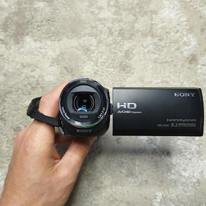 May include: A black Sony Handycam video camera featuring a Zeiss Vario-Tessar lens, HD AVCHD Progressive branding, and 9.2 Megapixels text on the side panel. The device includes a black wrist strap attached to the side of the lens housing.