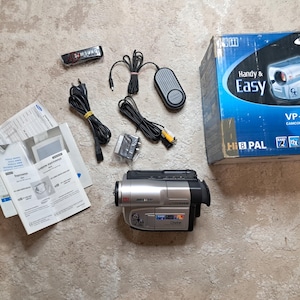 May include: A silver and black Samsung VP-L905D camcorder with accessories, including a remote, power cables, and instruction manuals. The camcorder is in its original box, which features the words "Handy & Easy".