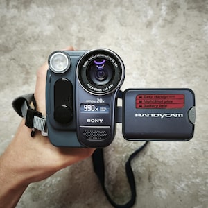 May include: A dark gray Sony Handycam video camera featuring a 20x optical zoom lens, a 990x digital zoom display, and a flip-out screen. The screen displays the text Easy Handycam, NightShot plus, and Battery Info in a red rectangular box.
