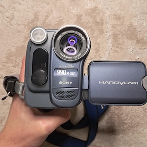 May include: A handheld Sony Handycam camcorder in shades of gray and blue. The camera has a lens with the text "OPTICAL 20X" and "SONY VIDEO LENS/OPTICAL 20X". The screen reads "990x DIGITAL ZOOM". The word "HANDYCAM" is printed on the flip-out screen.