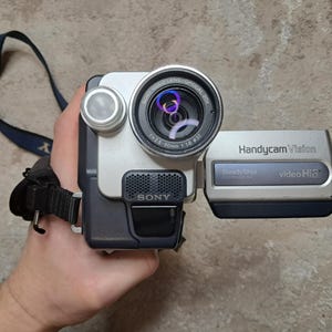 May include: Silver and black Sony Handycam Vision video camera. The camera has a lens with a silver ring, a microphone, and a flip-out screen. The camera has the words "Handycam Vision" and "video Hi8" printed on it.