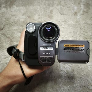 May include: A grey and black Sony Handycam camcorder featuring a 20x optical zoom lens, a 990x digital zoom display, and an open side panel. The panel displays text including Hi8/8mm Playback, Memory Stick Slot, and Touch Panel LCD.