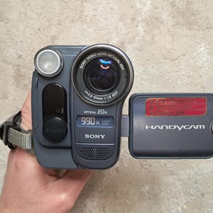 May include: A gray Sony Handycam camcorder with a black lens and a flip-out screen. The lens reads "Optical 20x" and "990x". The screen reads "Handycam". The camcorder is held by a hand.