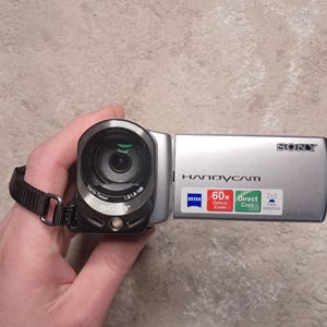 May include: A silver Sony Handycam camcorder with a black wrist strap. The camcorder has a Zeiss lens and features 60x optical zoom. The word "SONY" is printed on the side.