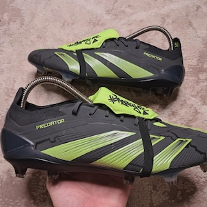 May include: Pair of gray and neon green soccer cleats with the word "PREDATOR" printed on the side. The cleats have a black strap and a neon green tongue with text. The cleats are on a neutral background.