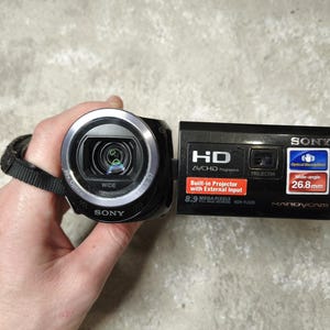 May include: A black Sony Handycam with a wide-angle lens, held by a hand. The camera has a built-in projector and features 8.9 megapixels. The text "Wide-angle 26.8mm" is visible on the side.