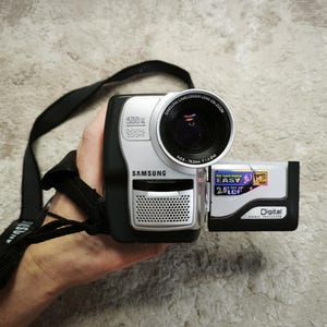 May include: A black and silver Samsung digital camcorder featuring a 500x digital zoom lens, a 2.5 inch LCD screen, and a black carrying strap. The device displays text including Samsung, 500x Digital Zoom, Easy, and Digital Signal Processor.