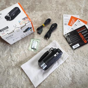 May include: A black Sony HDR-CX405 camcorder with its original white cardboard packaging box, two black USB cables, a small silver battery pack in a plastic bag, and several printed instruction manuals and product registration documents.