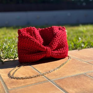 May include: A red crocheted handbag with a gold chain strap. The bag has a bow detail on the front.