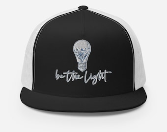 May include: A black and white trucker hat with a white mesh back. The front of the hat features a light bulb graphic with the text "be the light" in a stylized font.