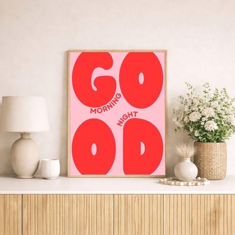 Op de afbeelding: Een rechthoekige wandprint met een lichtroze achtergrond en grote, dikke rode letters die het woord GOOD vormen. De woorden MORNING en NIGHT staan in kleinere rode letters tussen de letters gedrukt, alles in een dunne houten lijst.