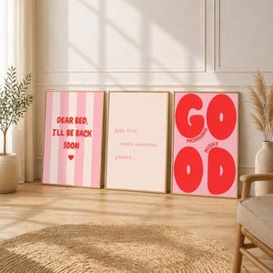 Op de afbeelding: Drie ingelijste wandposters. De linker poster heeft roze verticale strepen met de rode tekst 'DEAR BED, I'LL BE BACK SOON' en een hartje. De middelste poster is lichtroze met de tekst 'Just five more minutes, please...'. De rechter poster is roze met de rode tekst 'GOOD MORNING' en 'GOOD NIGHT'.