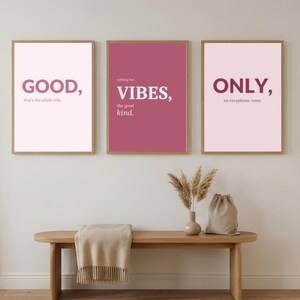 Könnte beinhalten: Drei gerahmte Kunstdrucke mit inspirierenden Zitaten. Die Drucke zeigen die Wörter "GOOD", "VIBES" und "ONLY" in einer modernen Schriftart. Die Farbpalette umfasst Rosa- und Burgundertöne. Vor den Drucken befindet sich eine Holzbank mit einer Decke, einer Vase und einer Tasche.