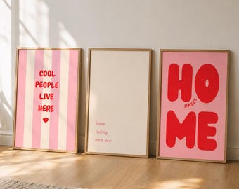 Op de afbeelding: Drie ingelijste rechthoekige wandposters. De linker poster heeft roze en crèmekleurige verticale strepen met rode tekst COOL PEOPLE LIVE HERE en een rood hartje. De middelste poster is crème met kleine rode tekst how lucky are we. De rechter poster is roze met grote rode tekst HO SWEET ME.