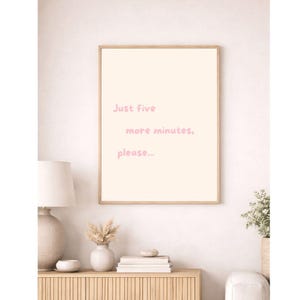 Op de afbeelding: Een rechthoekige wandkunstprint in een lichthouten lijst met een cr&egrave;mekleurige achtergrond en de tekst "Just five more minutes, please..." geschreven in een zacht, afgerond, lichtroze lettertype in het midden van de print.