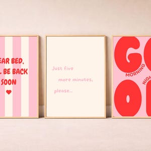 Op de afbeelding: Drie rechthoekige wandposters in lichte houten lijsten. De linker poster heeft roze strepen en de tekst "DEAR BED, I'LL BE BACK SOON" met een rood hartje. De middelste poster is cr&egrave;me met "Just five more minutes, please...". De rechter poster is roze met grote rode "GOOD" tekst en "MORNING" en "NIGHT" in kleinere rode letters.