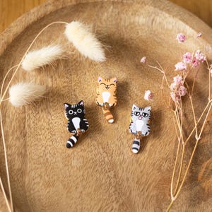 Könnte beinhalten: Drei Katzen-Emaille-Anstecknadeln in Schwarz-Weiß, Orange-Weiß und Grau-Weiß. Jede Anstecknadel hat einen hängenden Schwanz und liegt auf einem Holztablett mit getrockneten Blumen. Die Katzen haben unterschiedliche Gesichtsausdrücke.