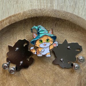 May include: A cute orange and white cat wearing a teal green witch hat with a pointed hat. The cat is wearing a white shirt and has a happy expression. The cat is standing on a silver pin back.