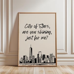 May include: A framed art print featuring a black and white skyline of a city, likely New York, with the text "City of Stars, are you shining, just for me?" in a handwritten font. The frame is a light wood color.