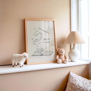 May include: A framed print with a light wood frame featuring a black and white illustration of a wand, book, and the text "You are so loved." A white lamb figurine and a teddy bear sit nearby. A white lamp with a pleated shade is also visible.