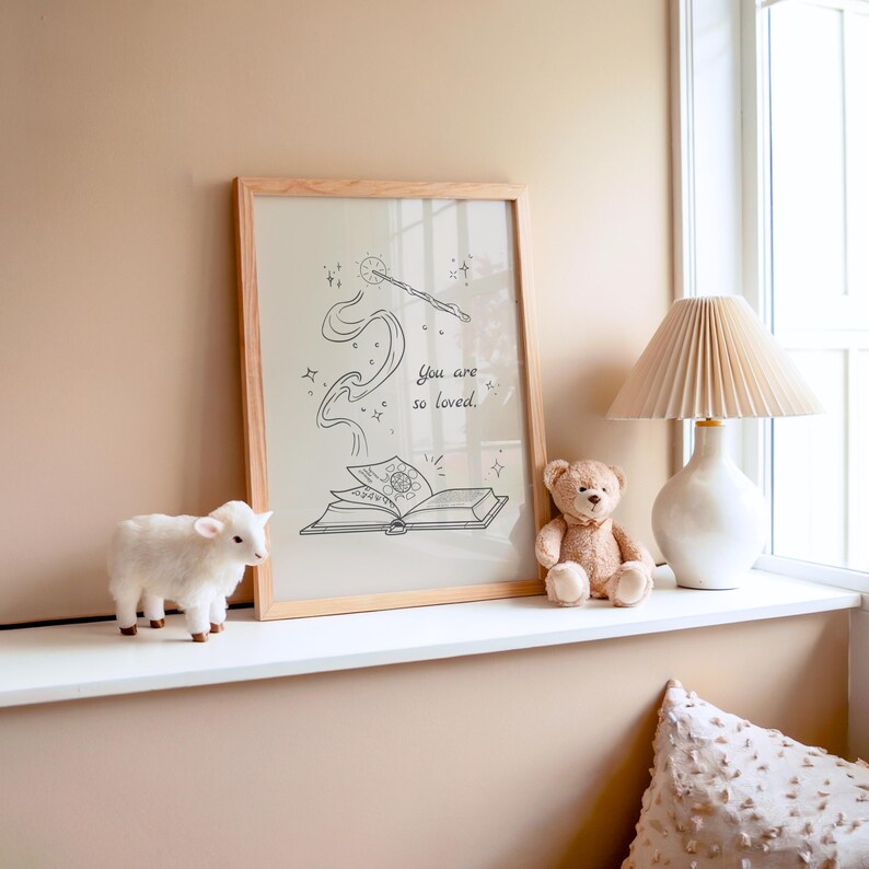 May include: A framed print with a light wood frame featuring a black and white illustration of a wand, book, and the text "You are so loved." A white lamb figurine and a teddy bear sit nearby. A white lamp with a pleated shade is also visible.