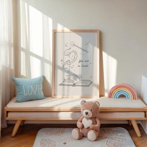 May include: A framed print with a whimsical illustration of a book and a wand, with the text "You are so loved." A light blue pillow with the word "LOVE" sits on a wooden bench. A teddy bear and a rainbow-shaped wooden toy are also present.