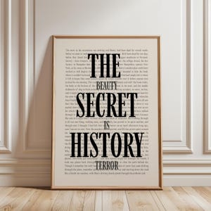 May include: Framed print with the words "THE BEAUTY SECRET IS HISTORY TERROR" in bold black letters. The text is layered over a background of printed book pages, creating a vintage aesthetic. The frame is a light brown color.