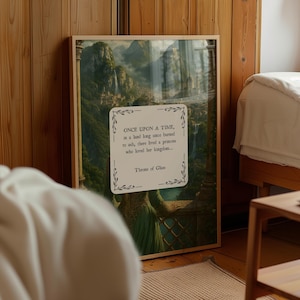 May include: A framed art print depicting a fantasy landscape with waterfalls and a mountain city. The print includes the text "ONCE UPON A TIME...Throne of Glass" within a decorative frame. The frame is a light brown colour.