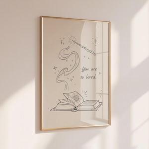 Puede incluir: Una impresión artística enmarcada con una ilustración caprichosa. La obra de arte representa un libro abierto con un diseño arremolinado, una varita mágica y el texto "You are so loved." La imagen es en blanco y negro con un marco dorado.