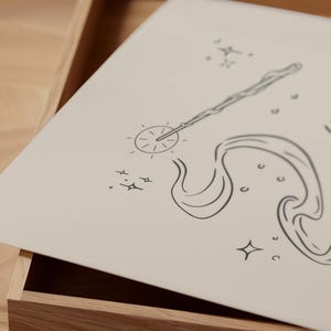 May include: A light beige drawing of a magic wand with a starburst design at the tip, and flowing lines. The drawing is on a light beige surface, with small star and circle accents. The drawing is inside a wooden box.