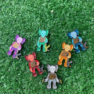 May include: A collection of seven colorful KAWS-inspired figurines, each holding a weapon. The figures are in various colors, including pink, green, blue, orange, red, and silver, with black accents. They are arranged on a green, grassy surface.