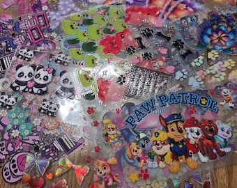May include: A collection of colorful, transparent stickers featuring various designs. The stickers include pandas, cherries, musical instruments, and characters from Paw Patrol. The stickers are arranged on a wooden surface.