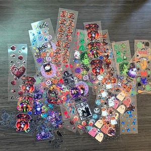 May include: A collection of Halloween-themed stickers on clear sheets. Designs include pumpkins, ghosts, witches, and the word "BOO". The stickers feature various colors and styles, perfect for crafting or decorating.
