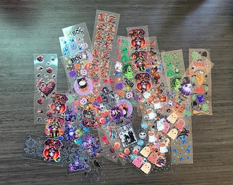 May include: A collection of Halloween-themed stickers on clear sheets. Designs include pumpkins, ghosts, witches, and the word "BOO". The stickers feature various colors and styles, perfect for crafting or decorating.