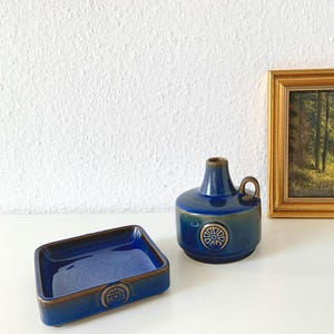 Könnte beinhalten: Ein Stillleben mit einer blauen Keramikschale und einer passenden Vase mit Henkel und dekorativem Emblem. Ein gerahmtes Landschaftsbild mit goldenem Rahmen ist ebenfalls vorhanden.