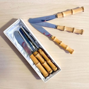 Könnte beinhalten: Ein Set Butterstreichmesser mit Edelstahlklingen und Bambus-Griffen. Die Messer werden in einer weißen Schachtel mit Satinfutter präsentiert. Die Griffe haben eine natürliche Holzfarbe und die Klingen sind silberfarben.