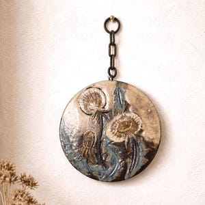Könnte beinhalten: Runde Keramik-Wandkunst mit einem Pusteblumen-Design in Beige-, Braun- und Blautönen. Aufgehängt an einer Metallkette und einem Haken. Ein dekoratives Element für die Inneneinrichtung.