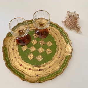 Könnte beinhalten: Ein rundes, grün-goldenes Tablett mit gewelltem Rand und einem zentralen Karomuster, begleitet von zwei klaren Weingläsern mit bernsteinfarbenen Stielen und Goldrändern sowie einer einzelnen strukturierten Muschel neben dem Tablett.