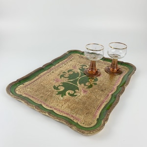 Könnte beinhalten: Ein rechteckiges Serviertablett mit gewelltem Rand, das ein abgenutztes Gold-Finish, grüne Ränder und rosa Akzente aufweist. Zwei klare Gläser mit bernsteinfarbenen, gerippten Stielen und goldfarbenen Rändern stehen auf der Oberfläche des Tabletts.