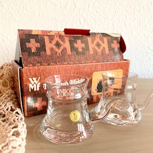 Könnte beinhalten: Zwei klare Kaffeetassen aus Glas mit Griffen, vor einer braunen Schachtel mit Muster. Die Tassen haben eine einzigartige Form und ein kleines goldenes Emblem. Die Schachtel hat das WMF-Logo.